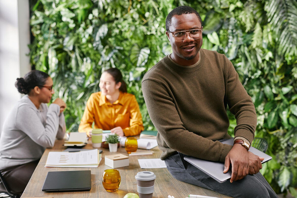 A man smiling at a meeting table with colleagues in a green, plant-filled workspace