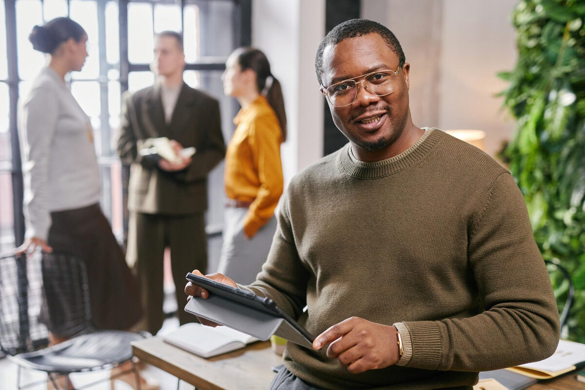 A man smiling while holding a digital tablet, with colleagues talking in the background.