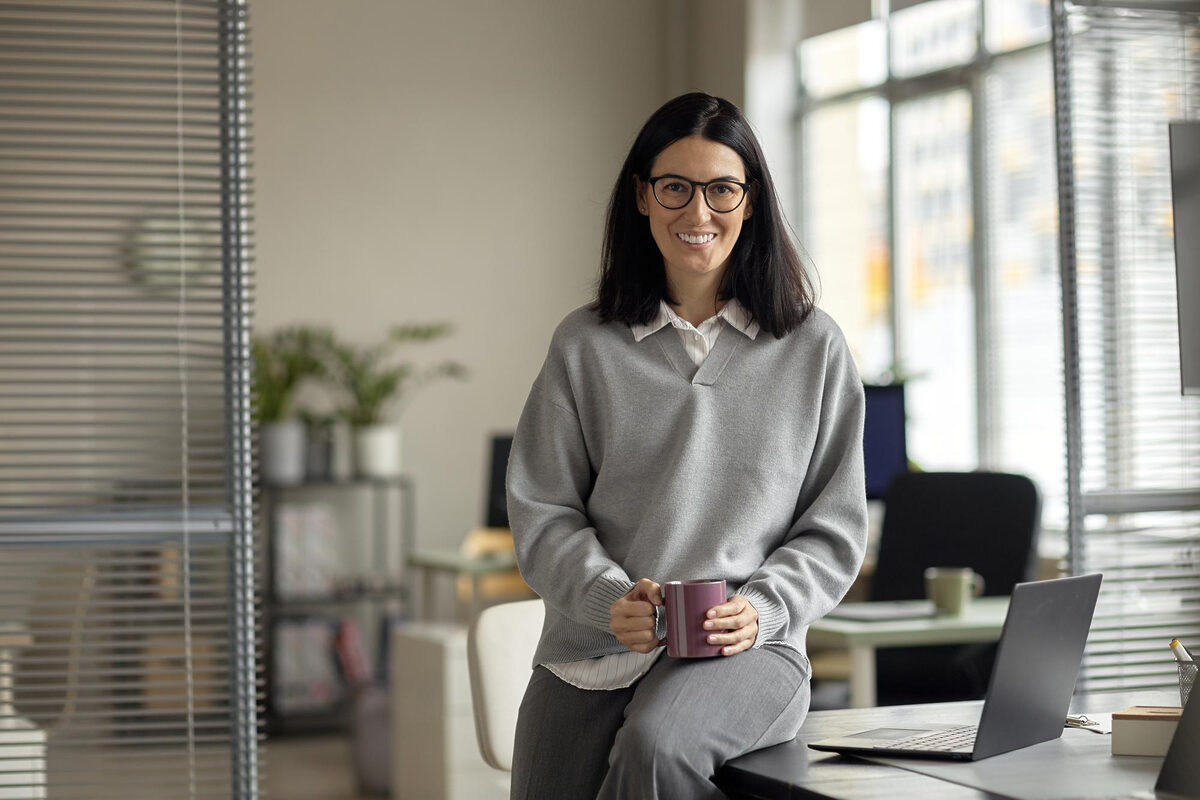 A woman sitting at a desk in a bright office, holding a coffee cup with a laptop nearby