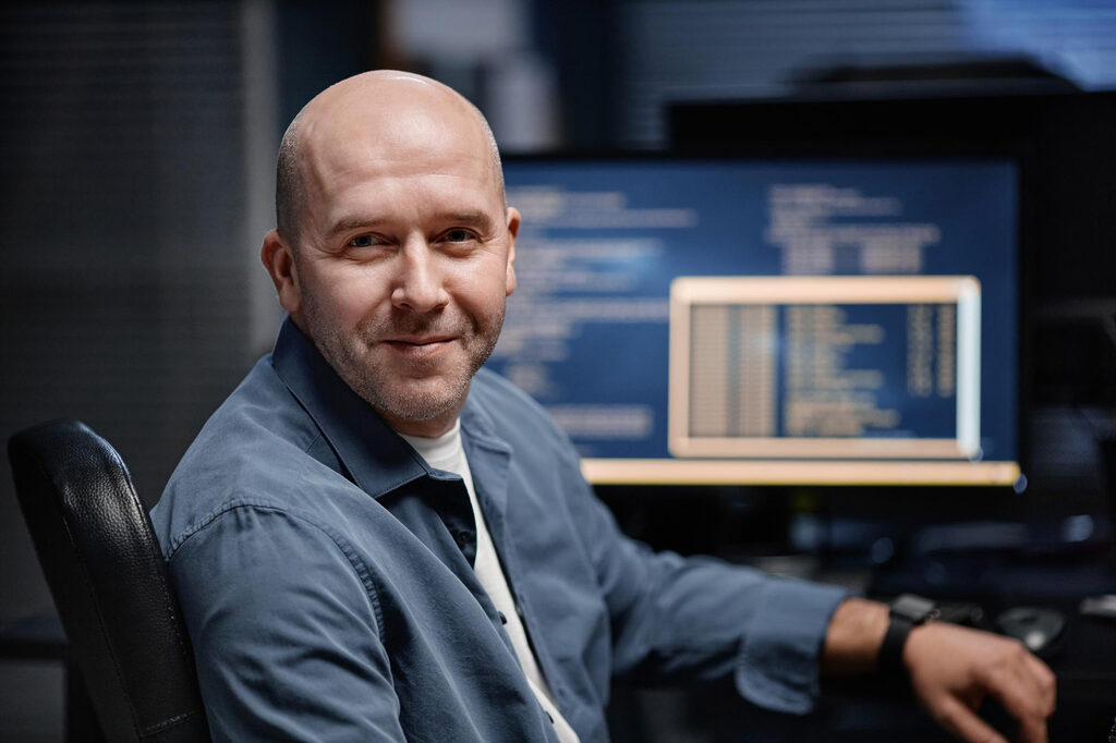 A man smiling in front of computer screens showing data graphs.
