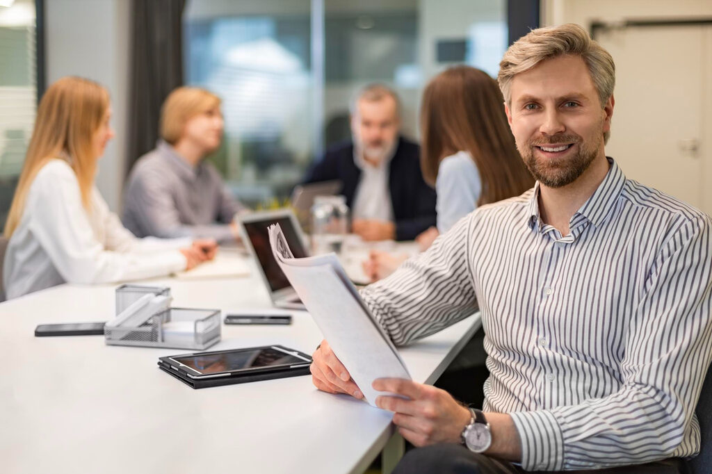 A man sitting in a meeting room holding a report with his team discussing